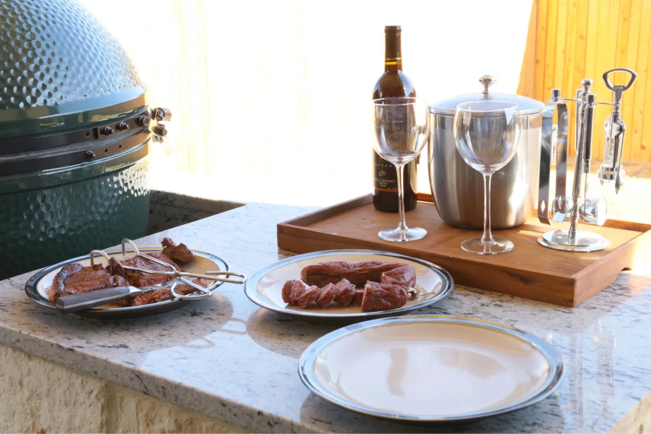 Outdoor grill setup with plates of meat, wine, and glasses on a countertop.