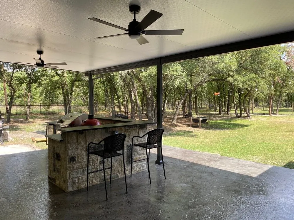 Covered patio with outdoor kitchen, two chairs, and ceiling fans, overlooking trees.