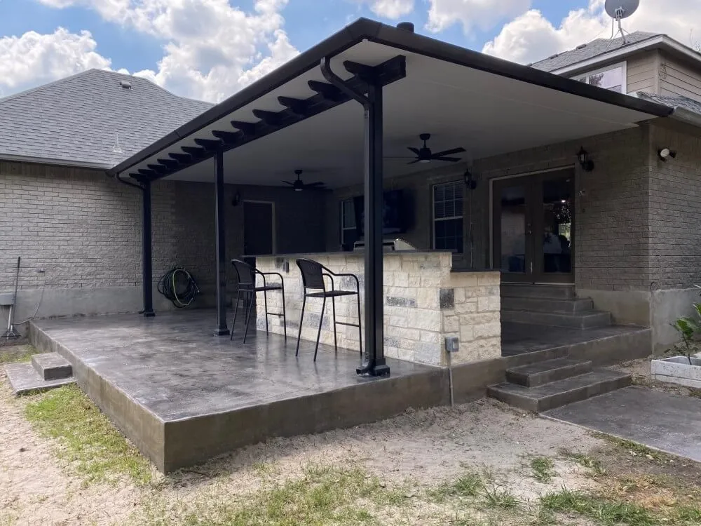 Modern backyard patio with covered seating area, bar stools, and stone bar under a cloudy sky.