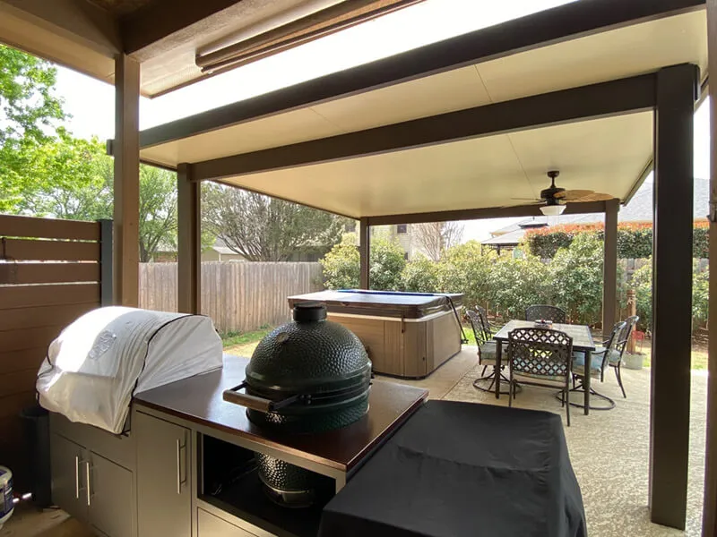 Covered patio with outdoor grill, hot tub, and dining table, surrounded by greenery and wooden fence.