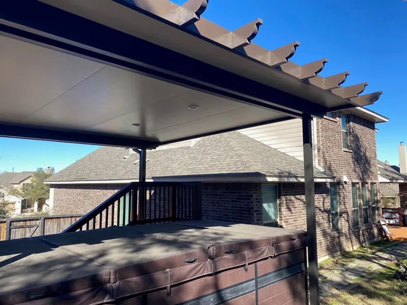 Covered outdoor patio with pergola above a hot tub, adjacent to a brick house under clear skies.