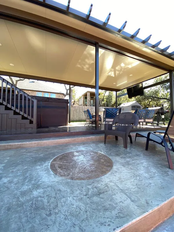 Covered patio with chairs, table, and decorative concrete floor, surrounded by trees and modern architecture.