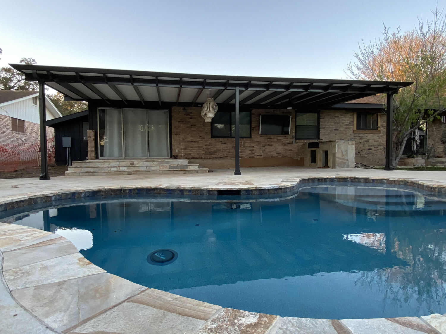 Modern backyard patio with a covered terrace overlooking a clear, blue swimming pool surrounded by stone tiles.
