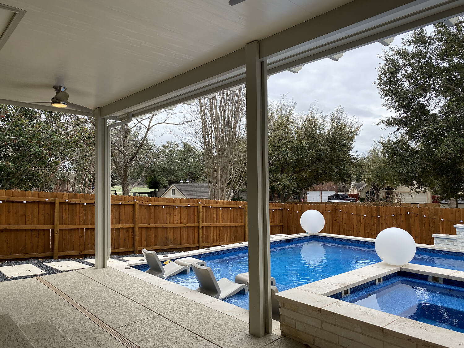 Covered patio overlooking a backyard pool with lounge chairs and decorative spheres, surrounded by a wooden fence.