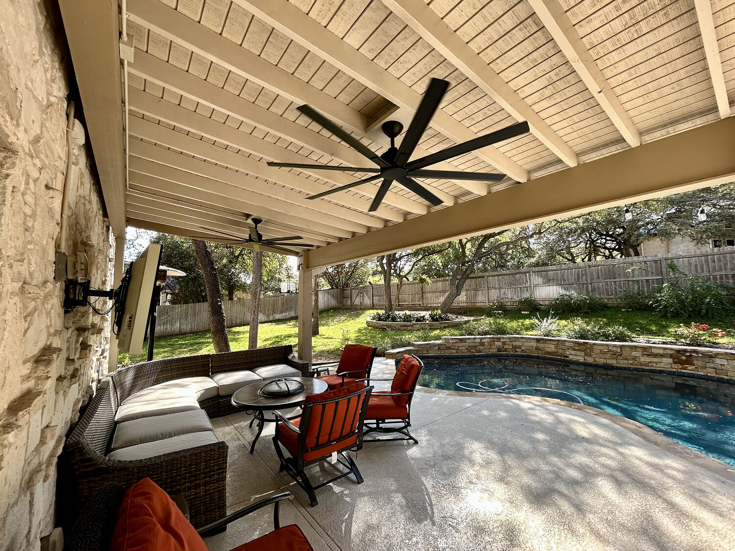 Covered patio with seating area and ceiling fan overlooks a backyard pool surrounded by greenery.