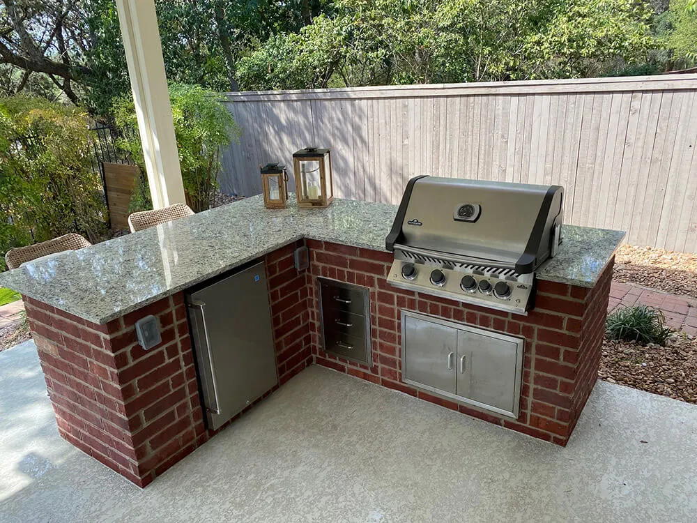Outdoor kitchen with brick island, granite countertop, grill, mini-fridge.