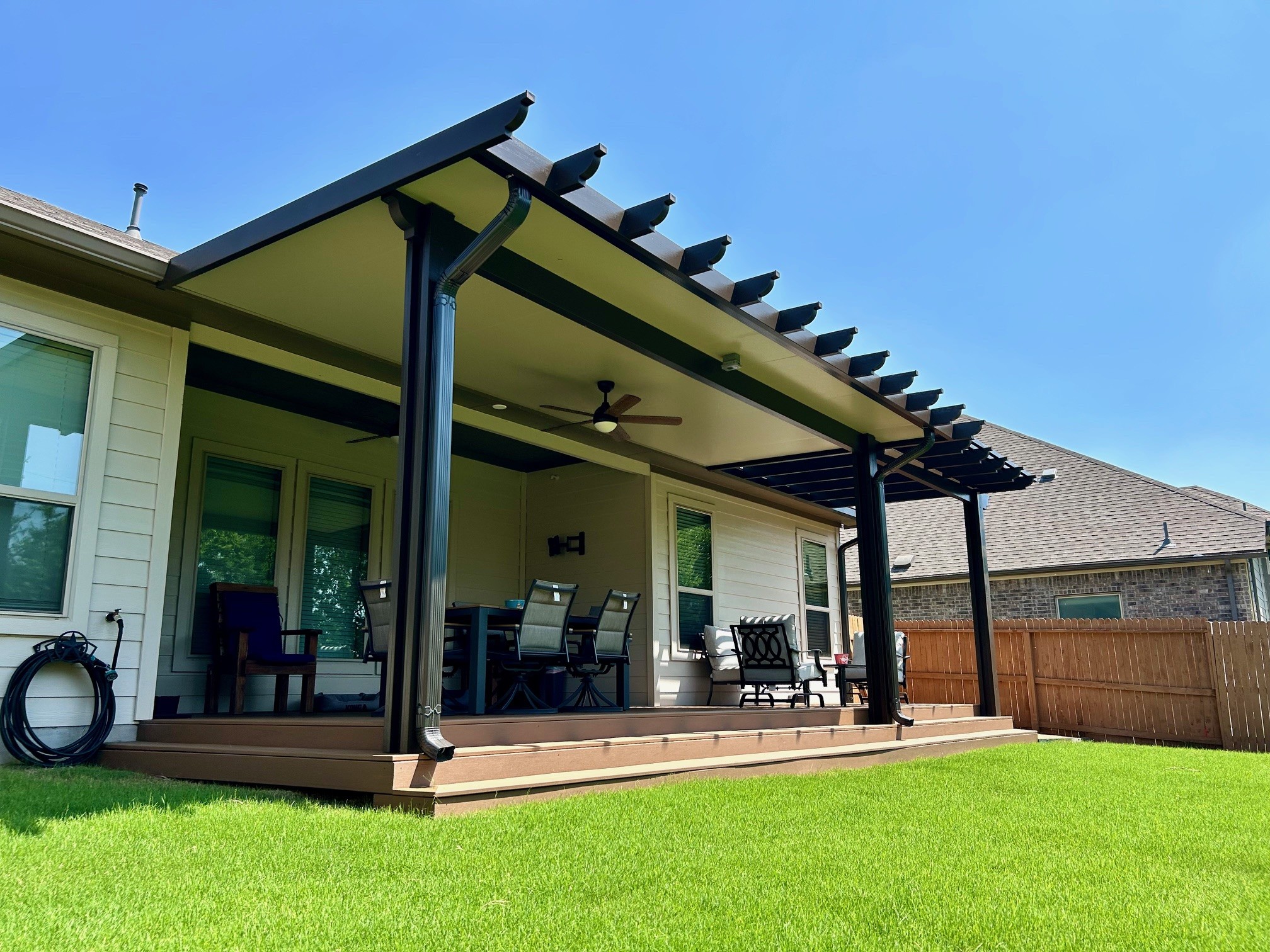 Backyard patio cover with attached pergola, outdoor furniture, and green lawn under a clear blue sky.