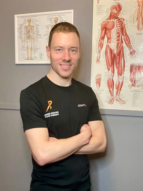 Smiling man with arms crossed wearing a black shirt standing in front of anatomical posters on a gray wall.
