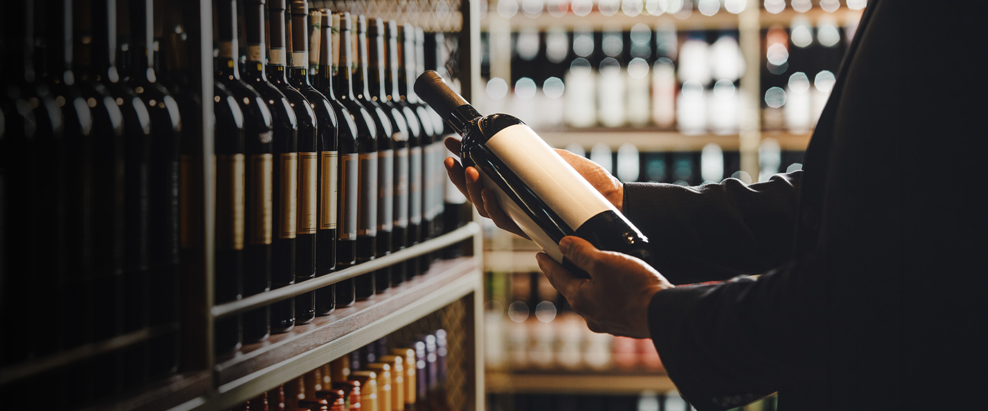 Person holding a wine bottle with blank label in a store aisle filled with wine bottles on the shelves.