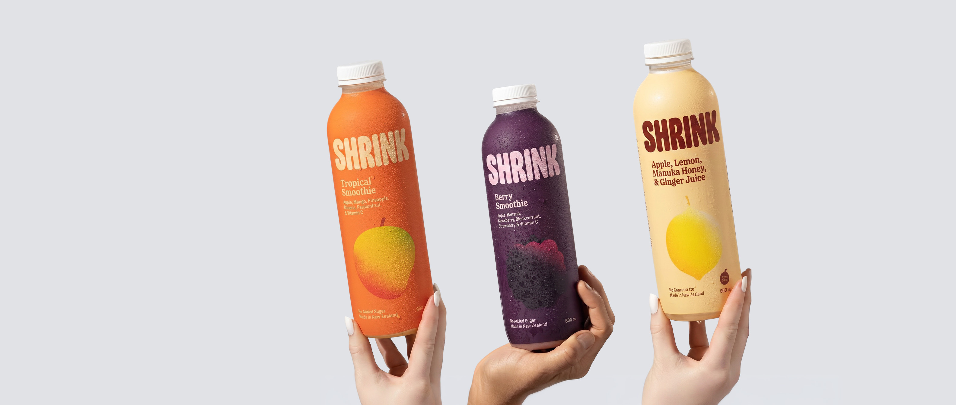 Three hands hold SHRINK smoothie bottles: tropical, berry, and apple-lemon ginger flavors with fruit images.