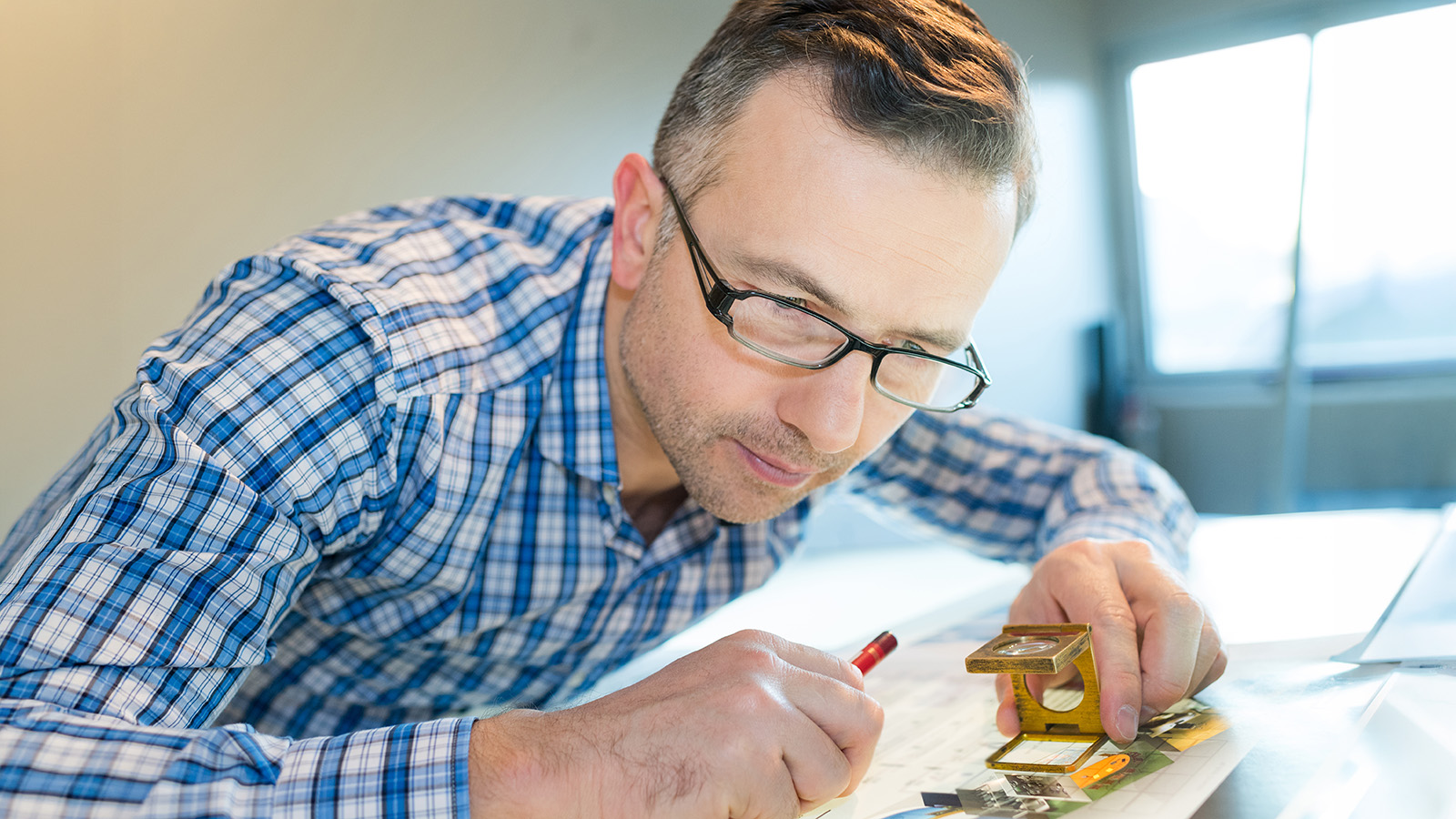 Man in glasses examining printed images on a table with a magnifier and red pencil.