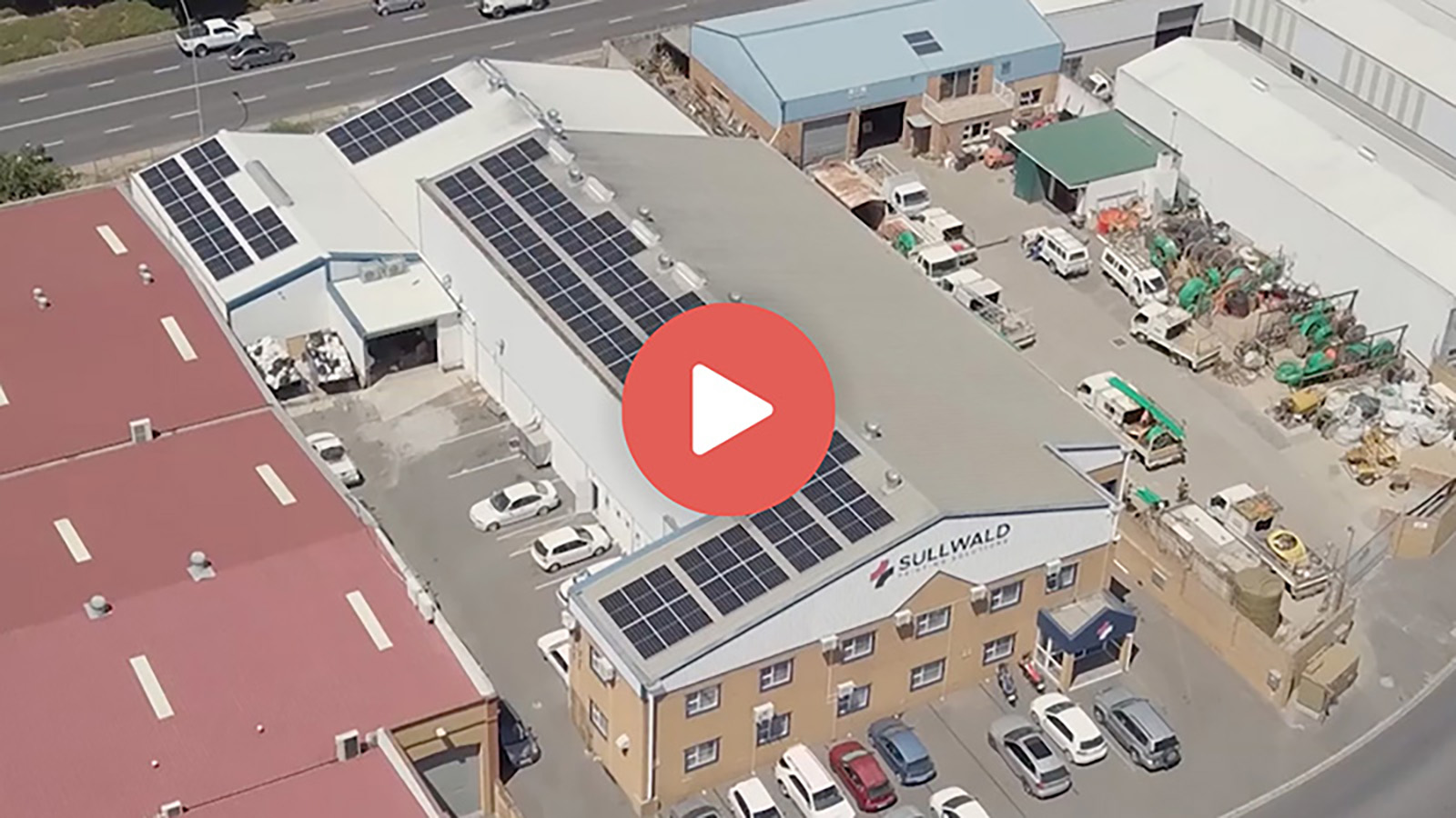 Aerial view of industrial buildings with solar panels on roofs and cars parked outside.