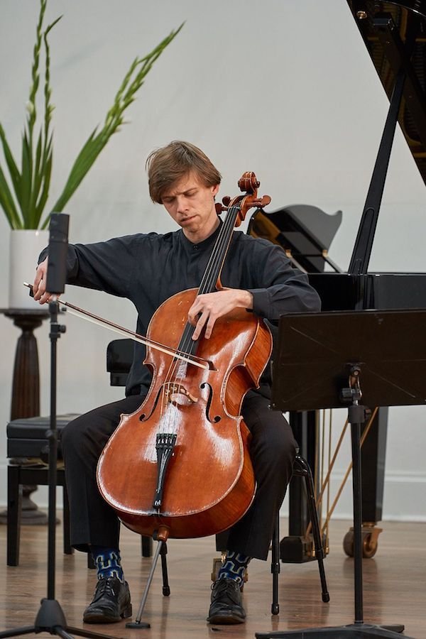 David Moran plays cello, in all black clothing, with patterned socks. There is a grand piano and a green plant behind him.
