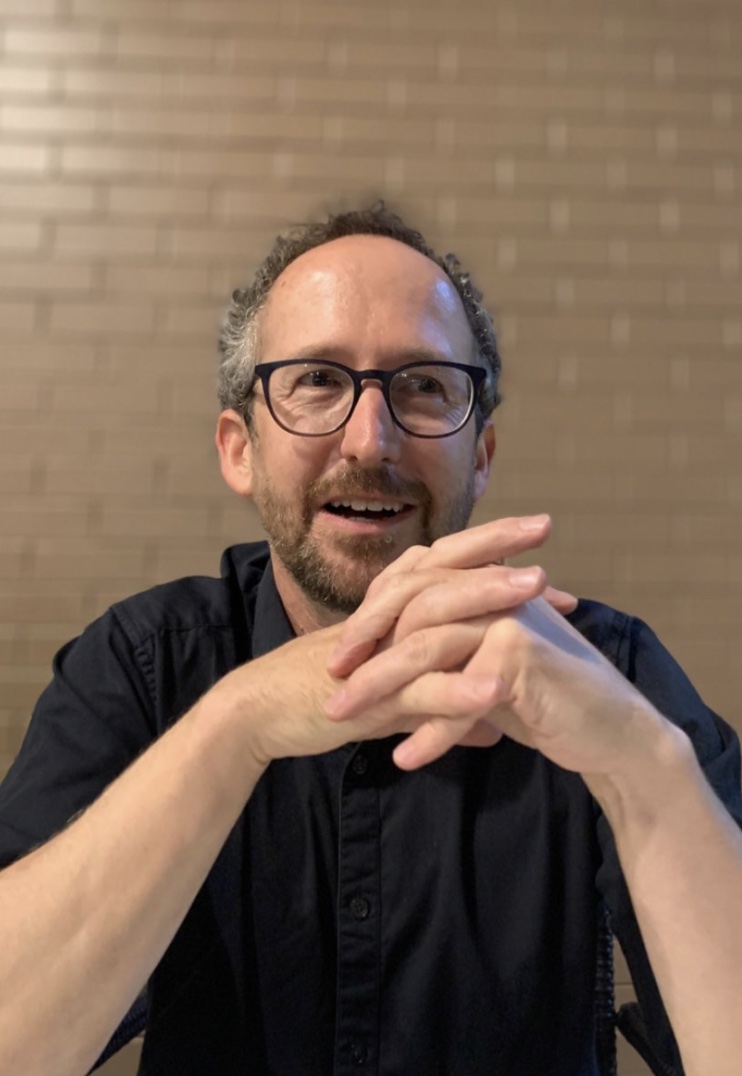 A close-up picture of Dave McEvoy. He has very short curly brown hair and glasses. He is smiling, with his elbows on the table and his hands clasped together in front of his chin.