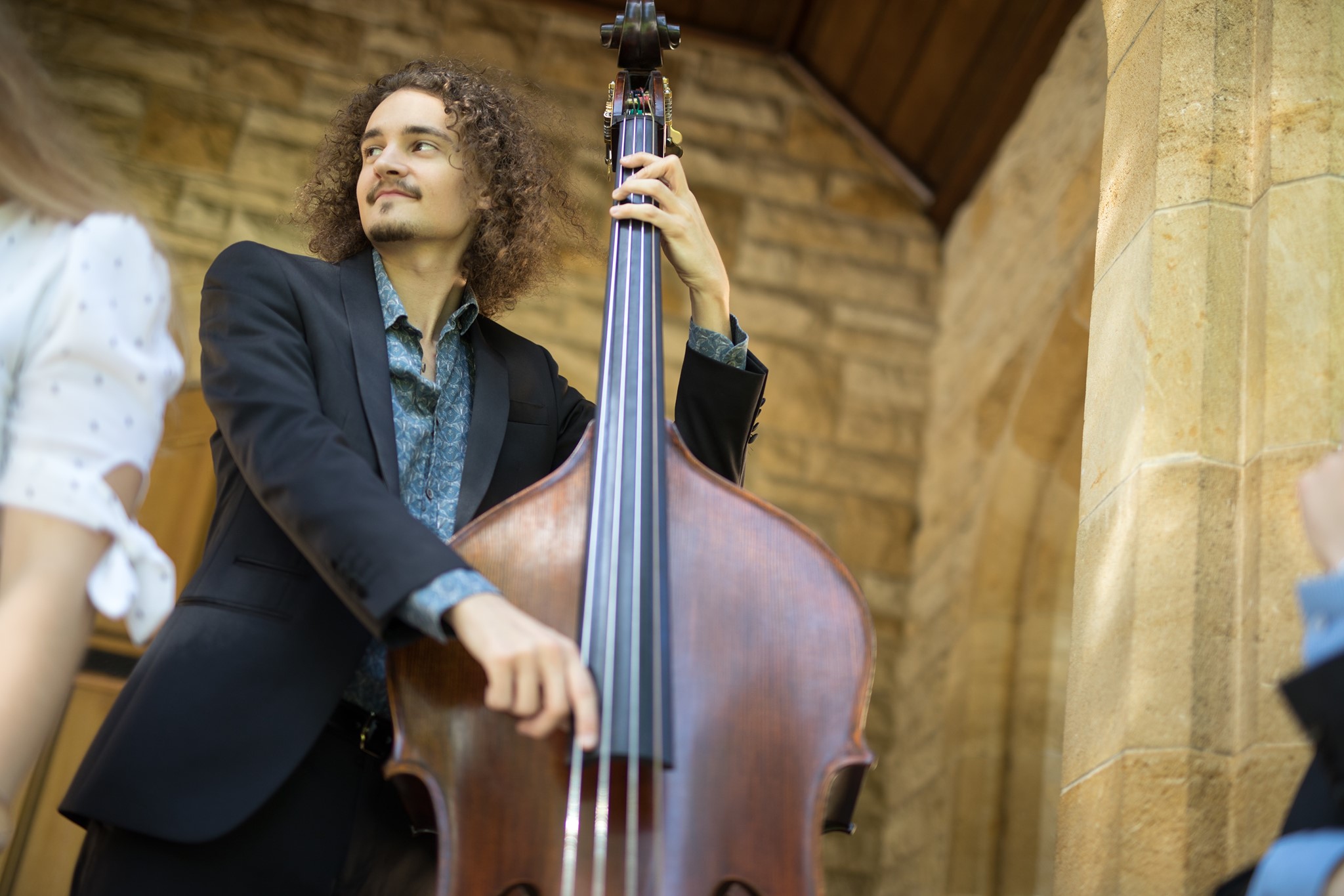 Dylan Paul plays double bass in front of a very old light brown stone wall, possibly in a church. He is wearing a navy blue suit. He has long curly brown hair and a moustache.