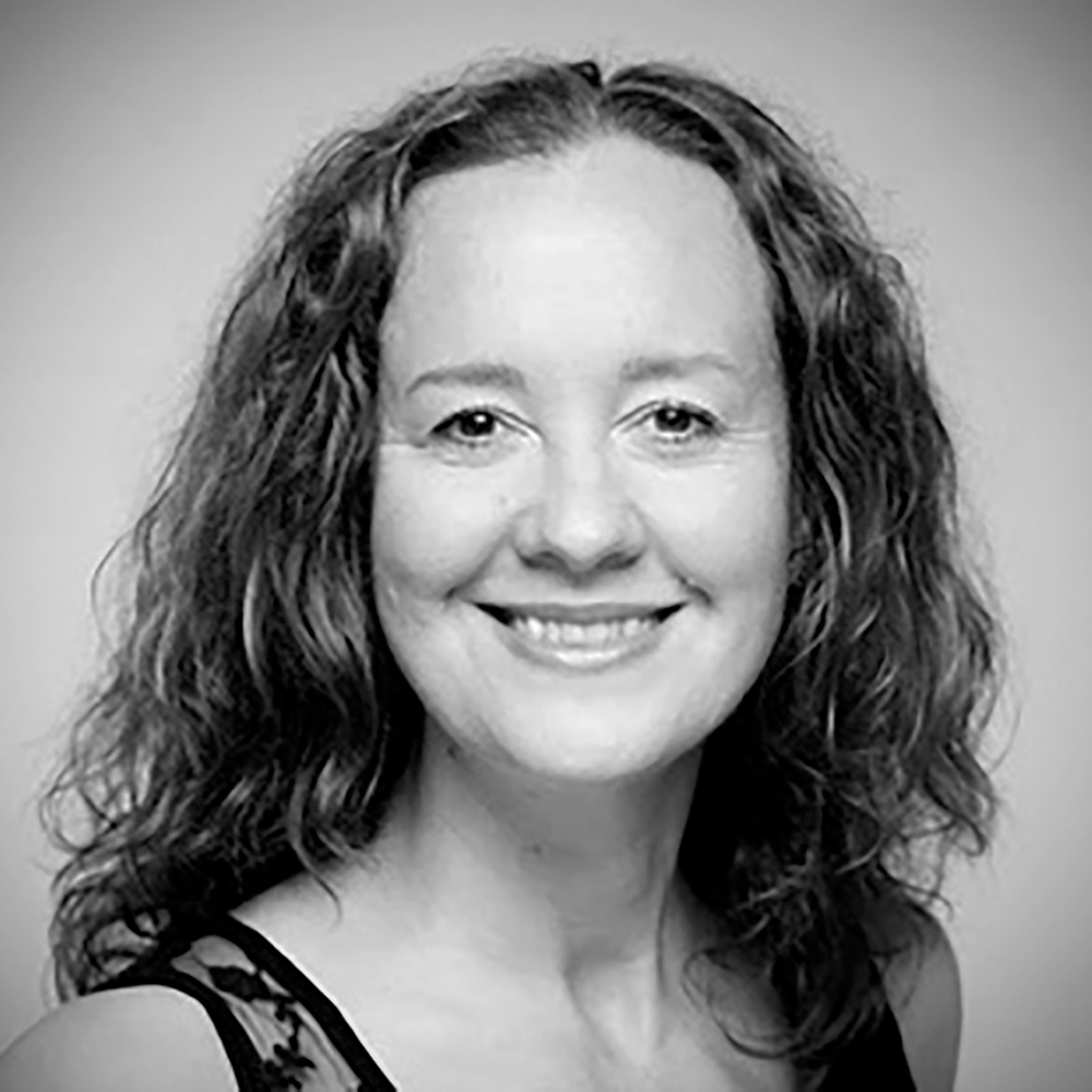 A close up, black & white headshot of Alison Rayner. She is smiling, and has wavy, shoulder-length brown hair.