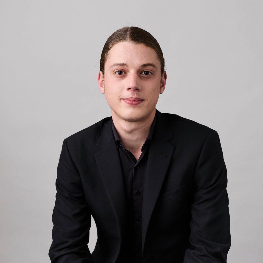 Justin Julian sits in front of a light grey background. He is wearing concert blacks. He has brown hair pulled back in a ponytail.