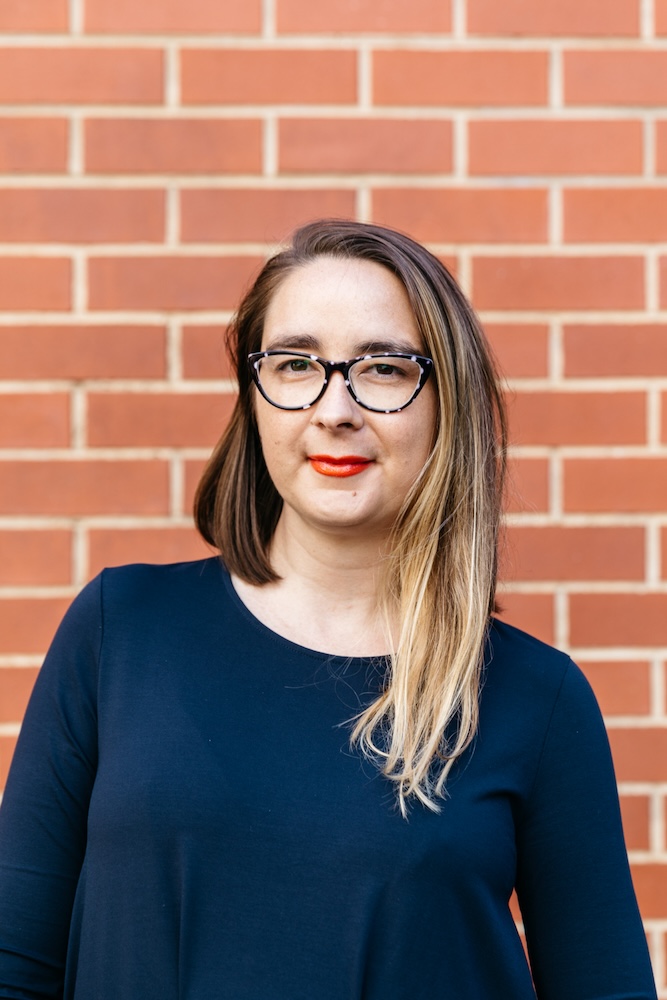 Emily Tulloch stands in front of a red-brick wall. She is wearing a long-sleeved navy shirt or dress. She has an asymmetrical haircut, very long on one side, brown with blonde towards the ends and shorter on the other. She has glasses and is smiling slightly.
