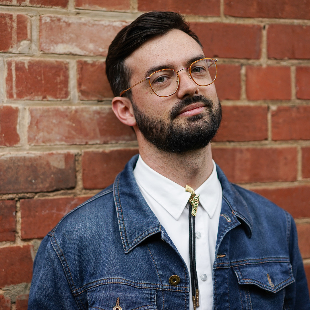 A photo of Kevin Van Der Zwwag from the chest up, in front of a brick wall. He has a white shirt, a denim jacket, and a bolo tie. He has brown hair and a beard, and is wearing glasses and smiling.