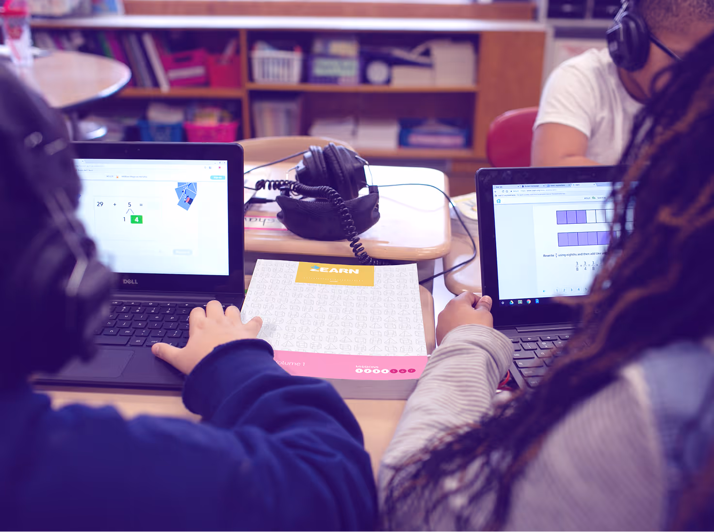 Two students wearing headphones using laptops at a desk with an educational workbook and headphones between them.
