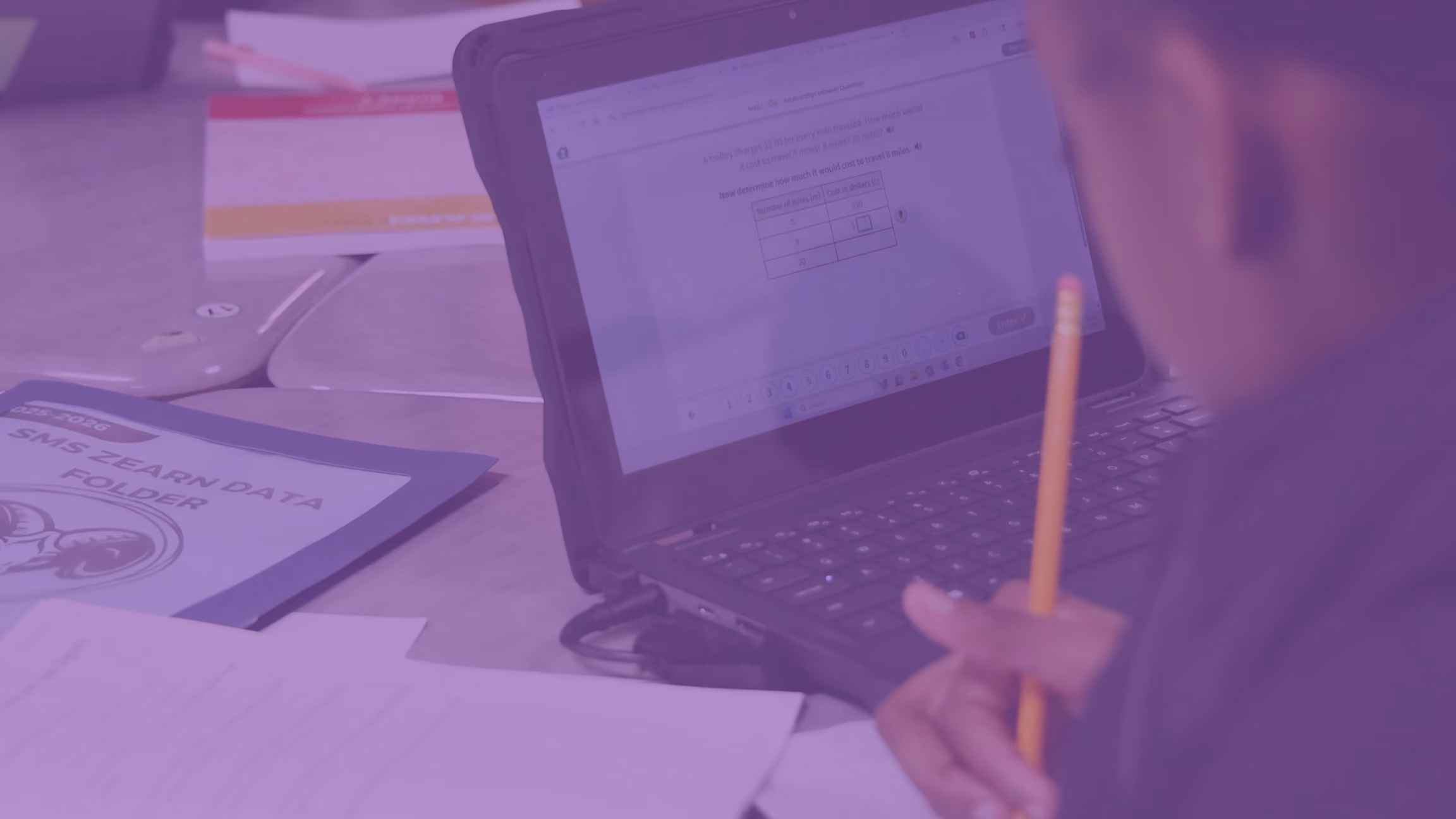 Student holding pencil, working on math problem on a laptop at a desk with papers and folder.