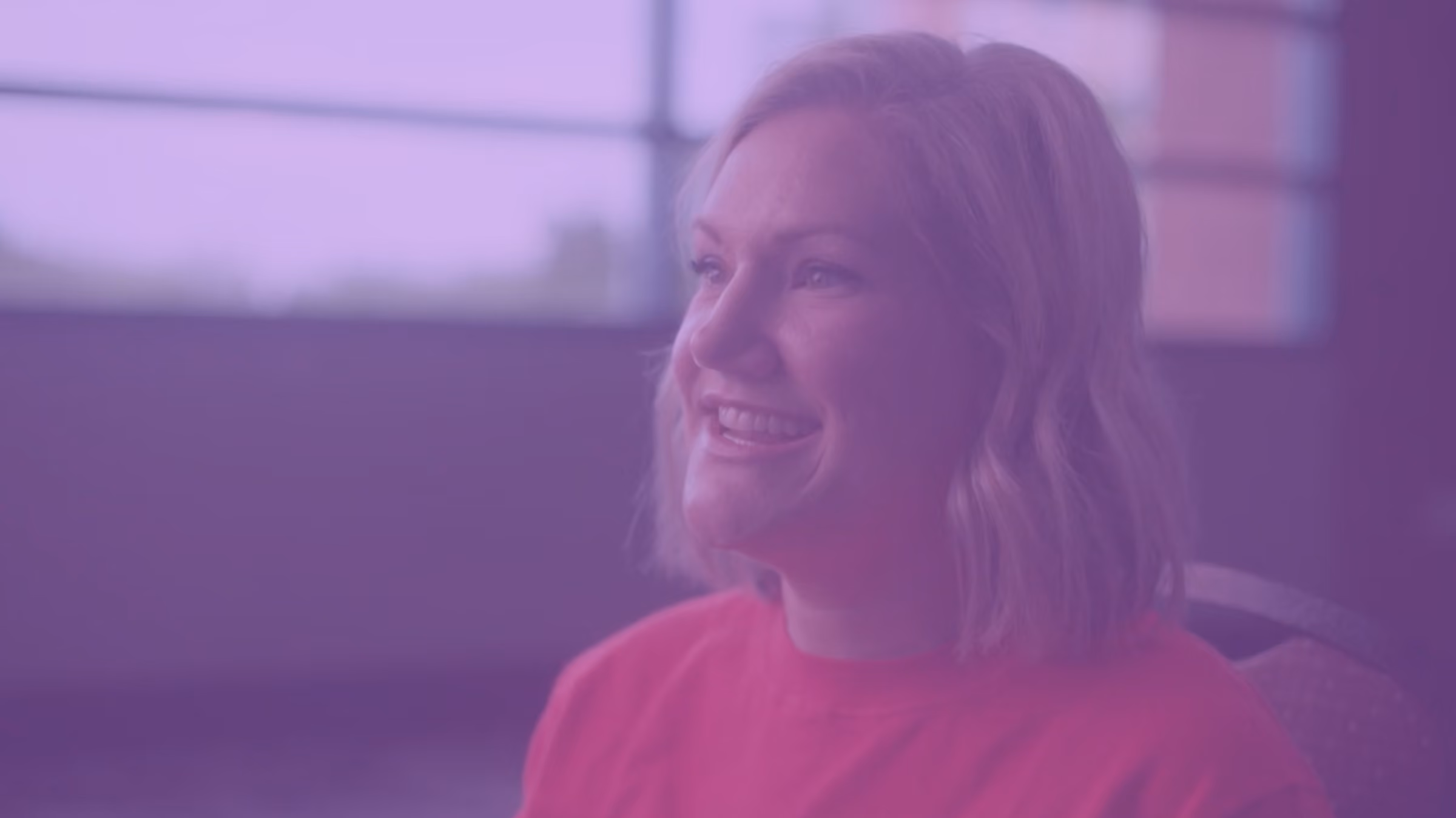 Smiling woman with short blonde hair wearing a red shirt sitting indoors near a window.
