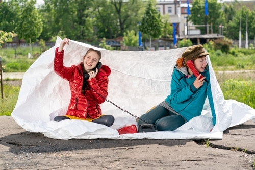 Zwei weibliche Personen unterschiedlichen Alters telefonieren an Wählscheibentelefonen im öffentlichen Raum auf weißer Decke sitzend.