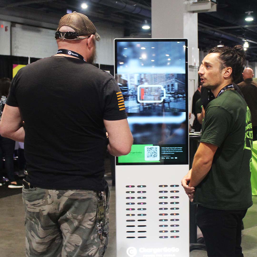 Two men stand by a charging station with a low battery icon on its screen indoors.