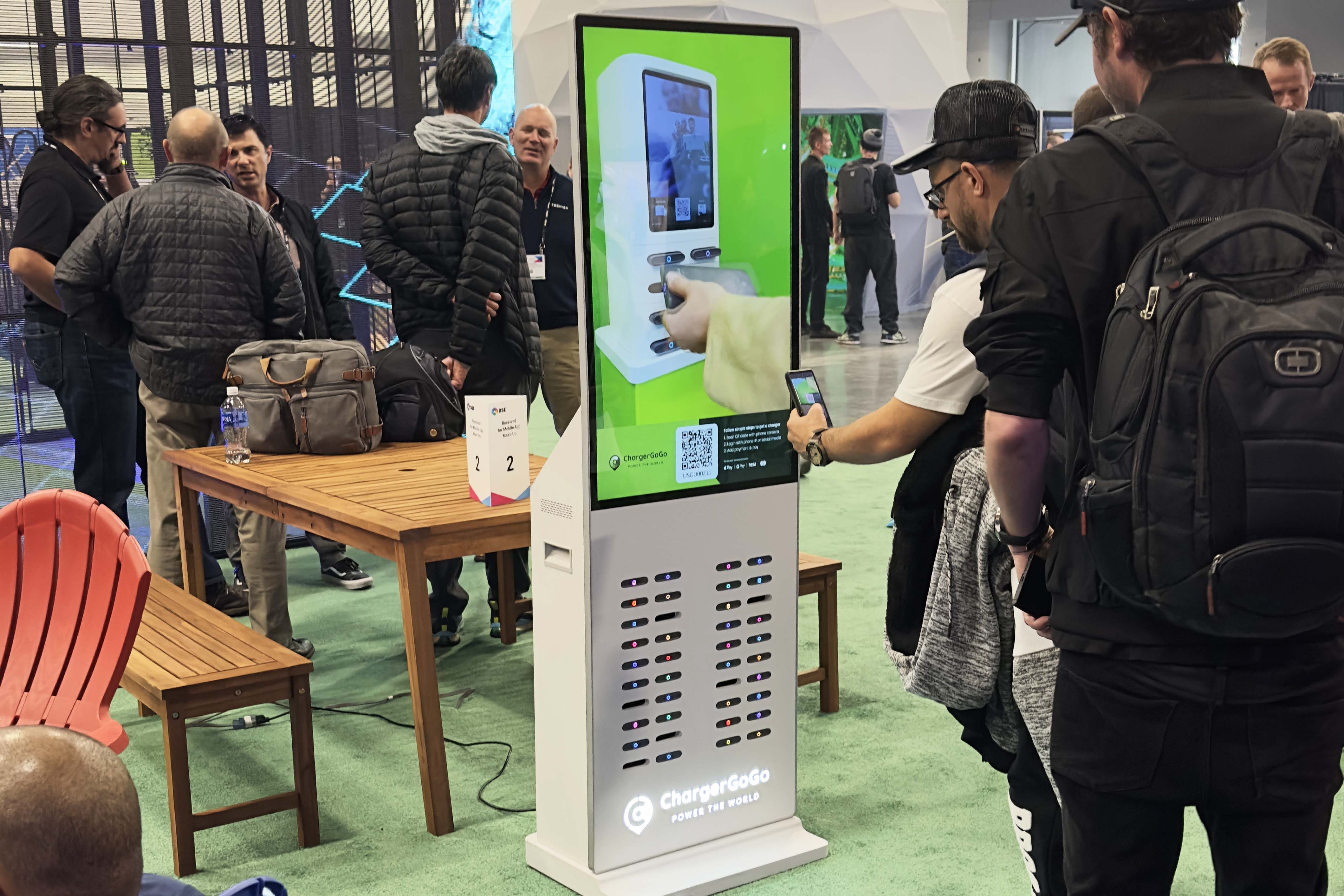 People interacting with tall white charging stations at an indoor exhibition or trade show.