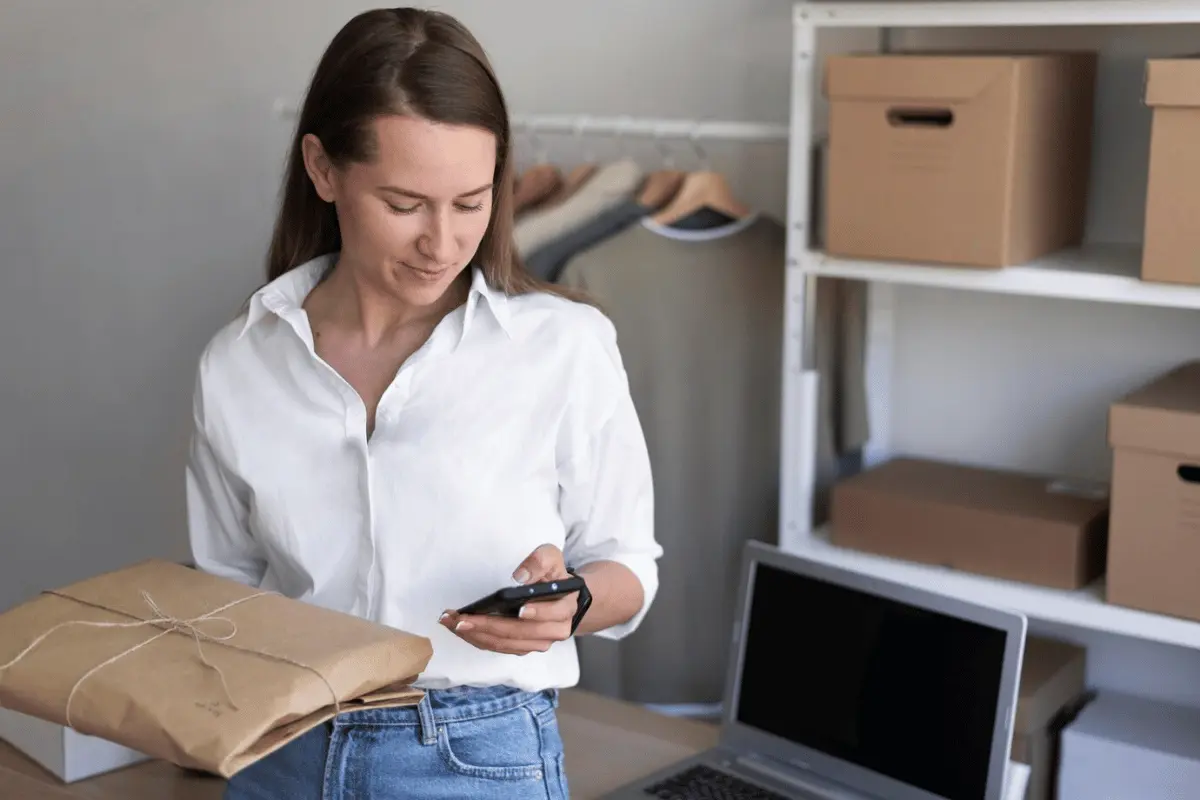 A person standing in front of a desk holding a package and looking at their cell phone.