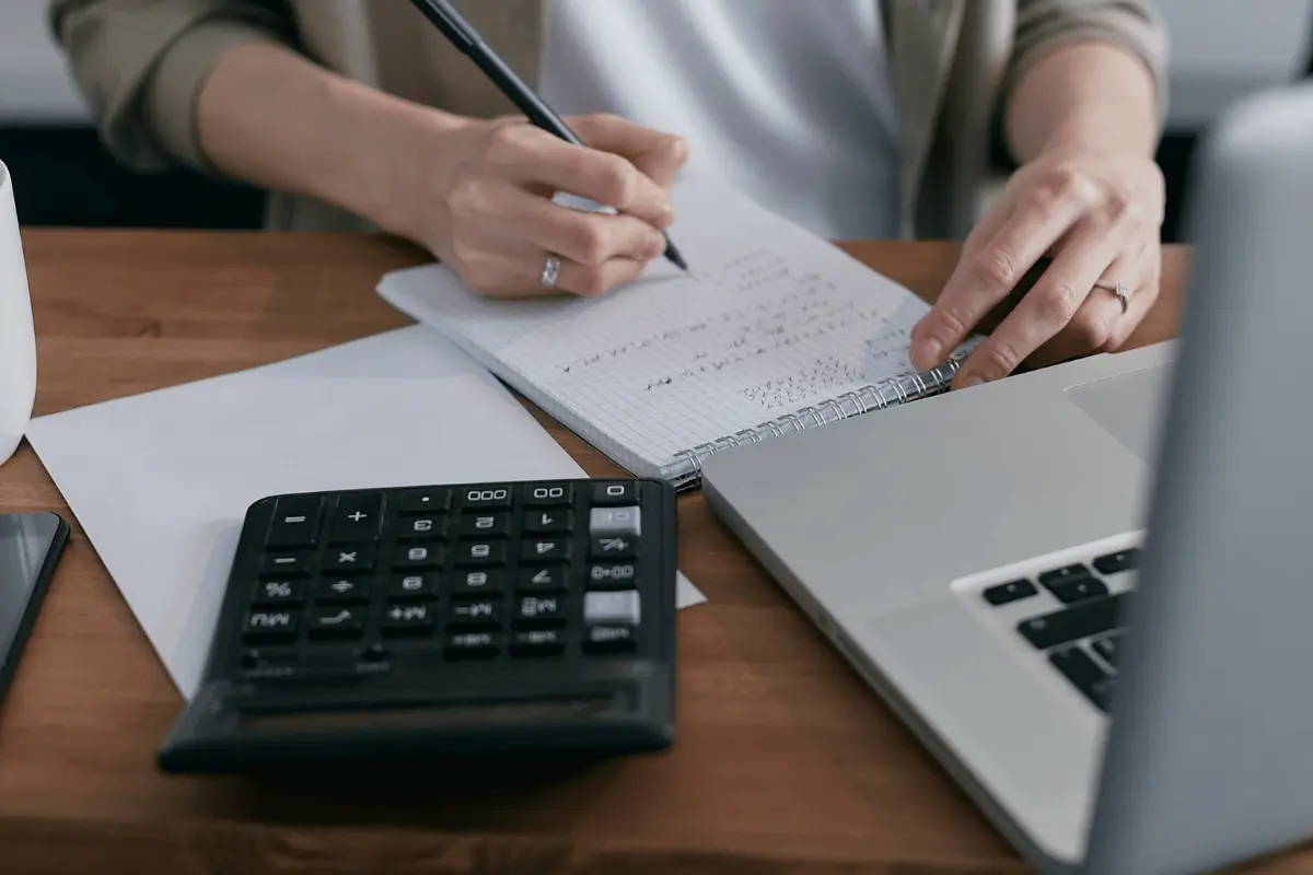 Person writing notes beside a laptop and calculator on a wooden desk.