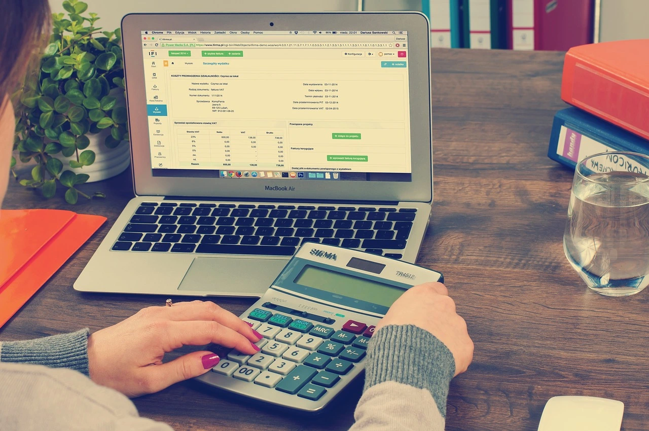 A woman holding a calculator in front of a silver laptop doing bookkeeping on there.