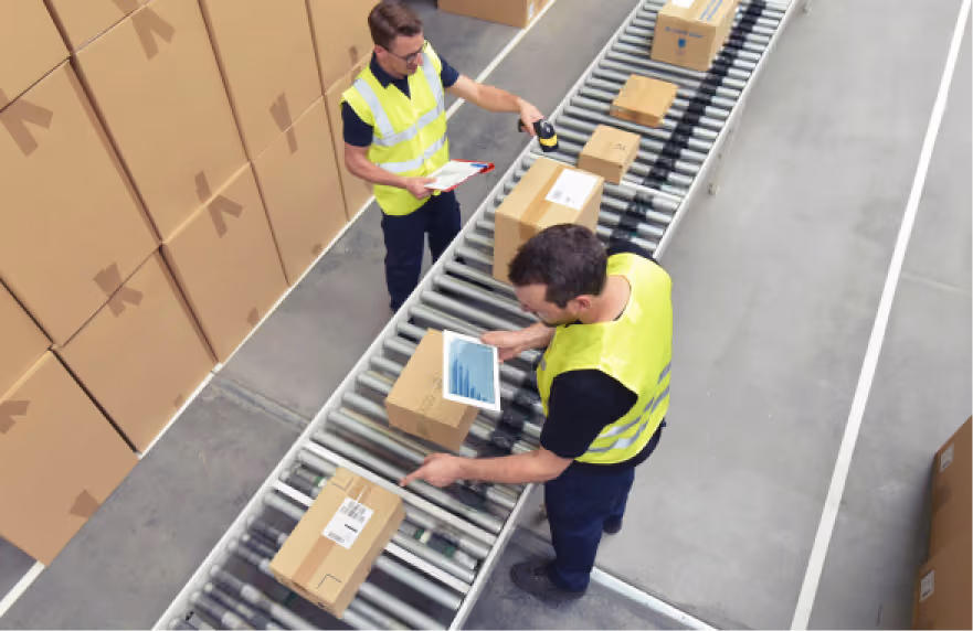 Two warehouse workers in safety vests process packages on a conveyor belt; one scans a box while the other checks information on a clipboard.