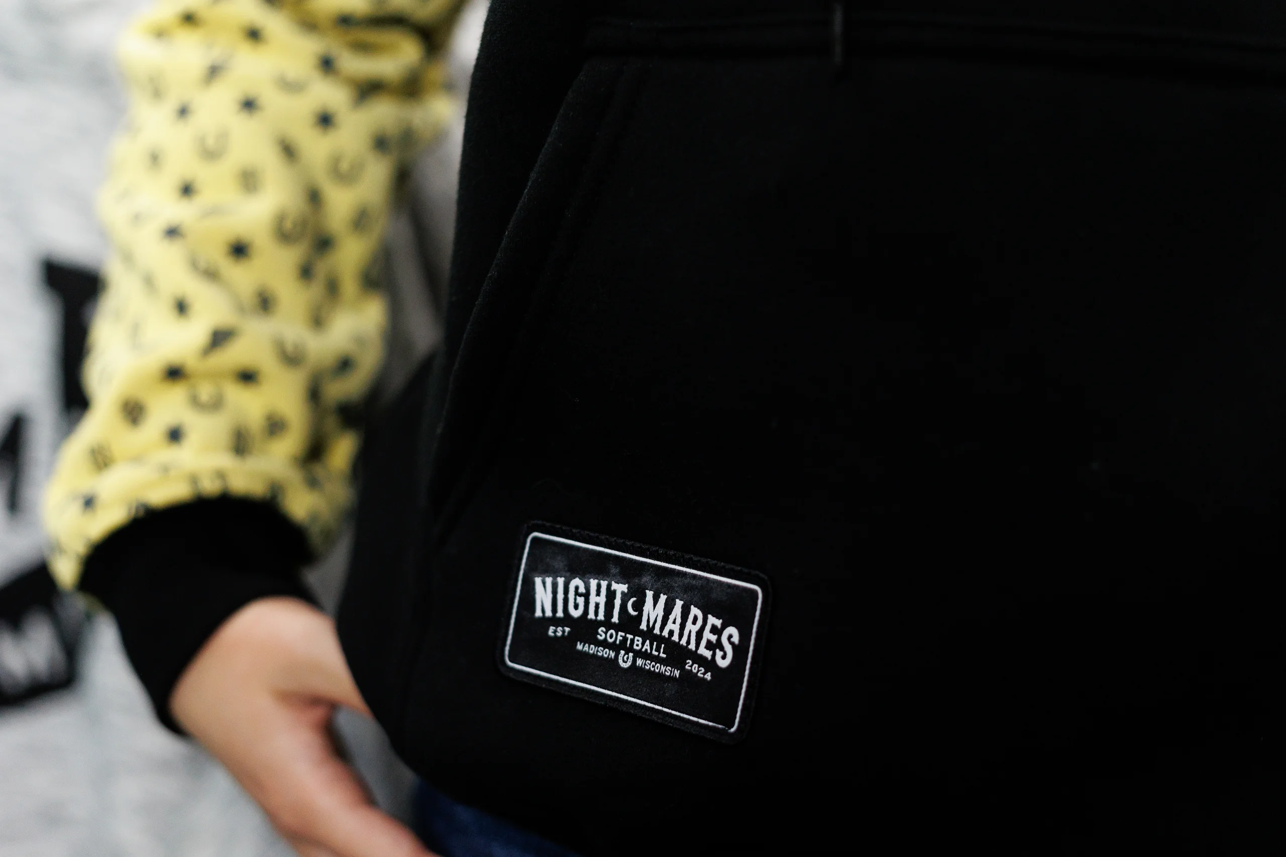 Close-up of a black hoodie pocket with a white patch reading 'NIGHT•MARES SOFTBALL EST 2024 MADISON WISCONSIN', worn by a person in a yellow patterned sleeve.