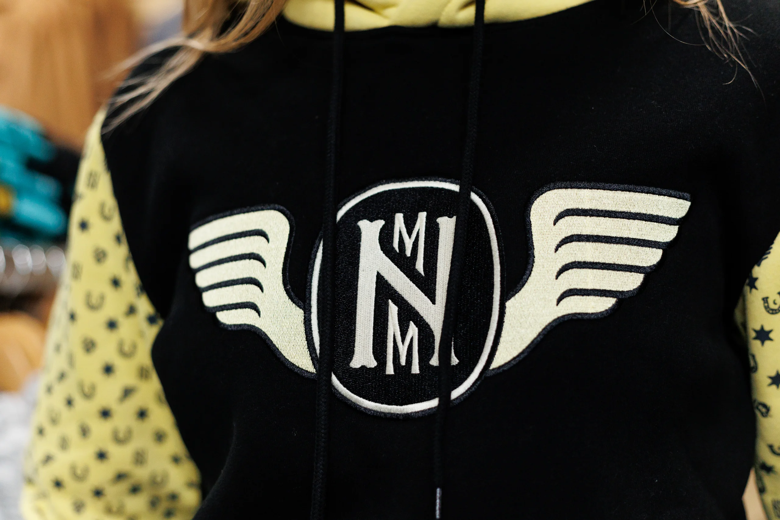 Black hoodie with a yellow winged emblem featuring the letters 'NMM' on the front and yellow sleeves patterned with small black icons.