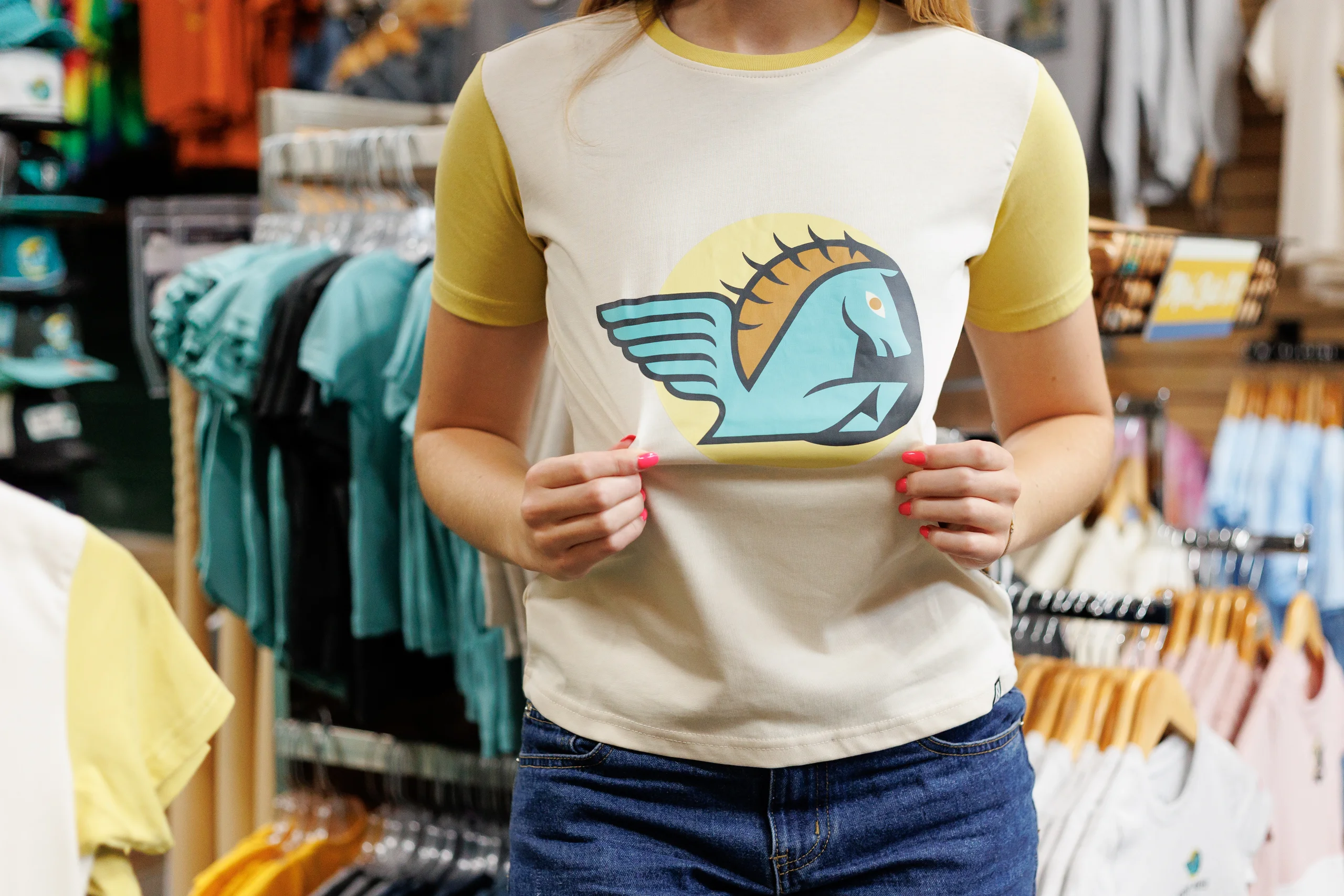 Person wearing a short-sleeve t-shirt with yellow sleeves and a graphic of a blue winged horse on a yellow circle in the center, standing in a clothing store.