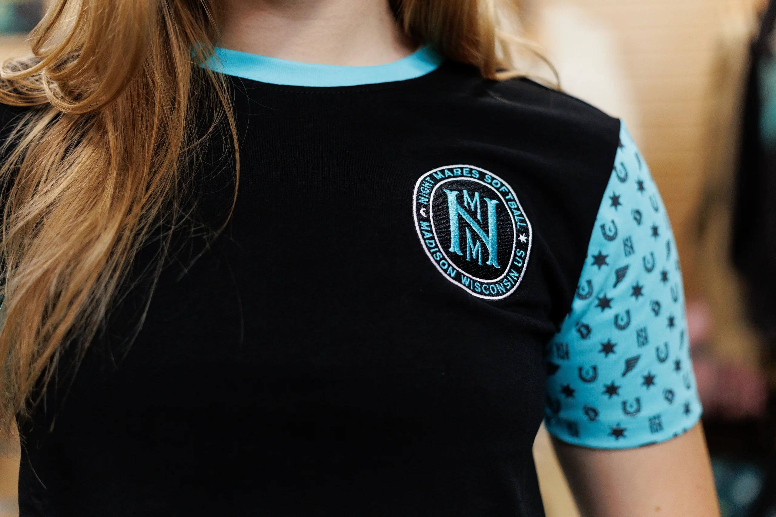 Close-up of a person wearing a black t-shirt with a light blue round collar, light blue sleeve with black symbols, and a circular patch reading 'Nightmares Softball Madison Wisconsin US.'