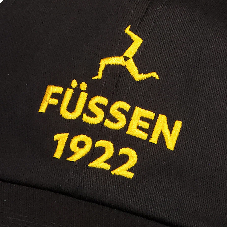 Close-up of black fabric with yellow embroidered text 'FÜSSEN 1922' and a yellow triskelion symbol above it.