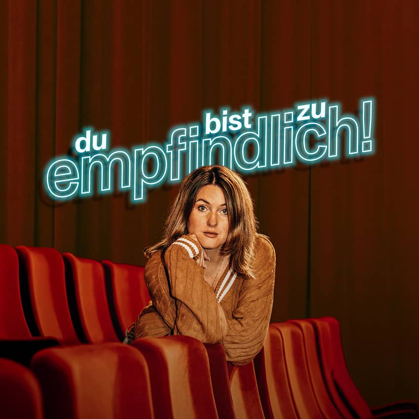 Kathi Kattinger im braunen Strickpullover sitzt in einem roten Theatersessel und stützt das Kinn auf die Hand, darüber Neonschriftzug „du bist zu empfindlich!“