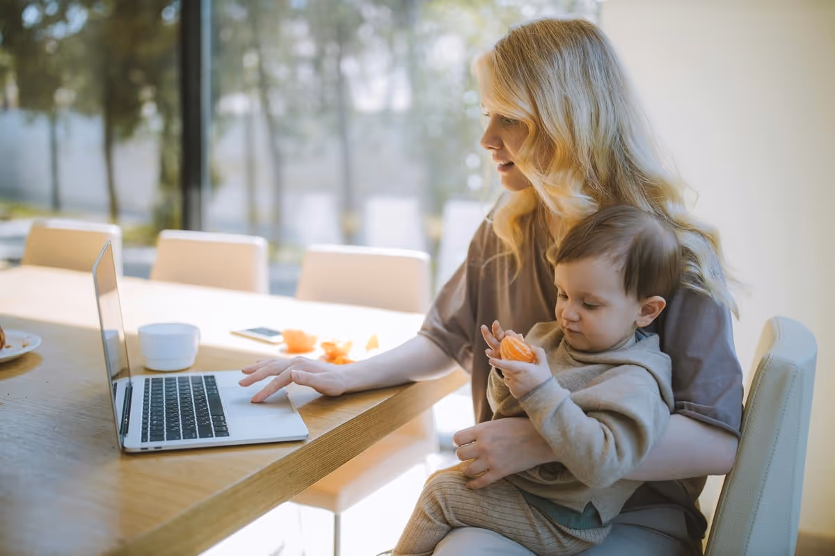 mother working on a laptop on a table holding kind on the lap. 