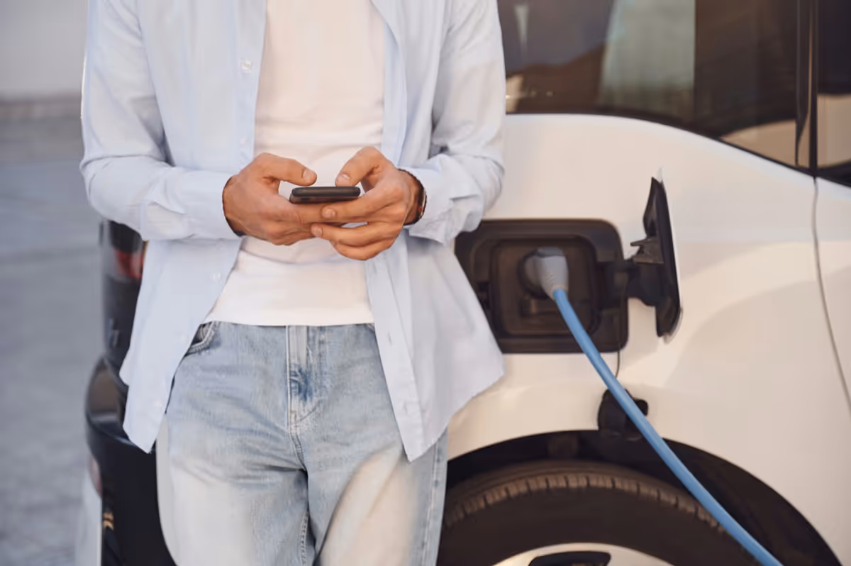 Person wearing light blue shirt and jeans using a smartphone while standing next to an electric vehicle being charged.