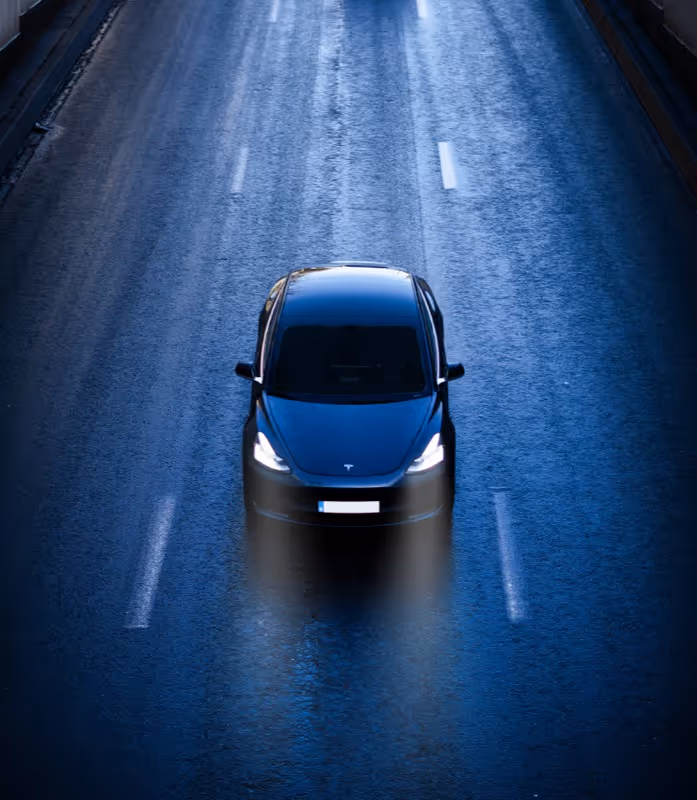 Black Tesla electric car driving on a wet road at night with headlights on.
