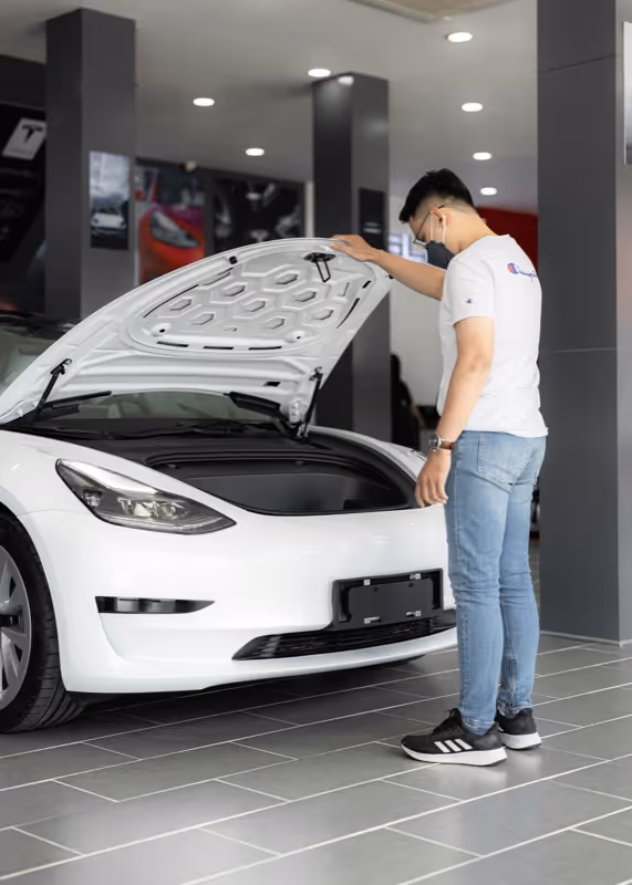 Man wearing mask inspecting the front trunk of a white electric car inside a showroom.