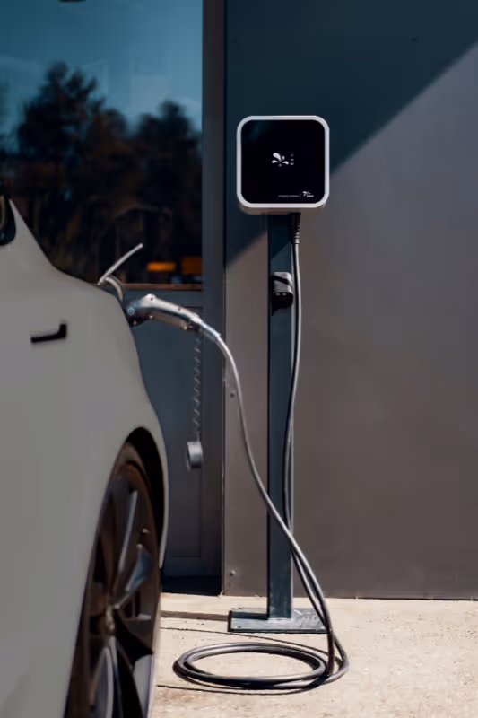 White electric car connected to a black wall-mounted charging station with a coiled cable.