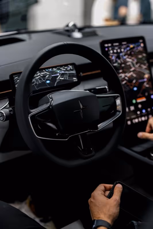 Interior of a car focusing on a black steering wheel with a digital dashboard and a large touchscreen displaying navigation maps, a person holding a smartphone is visible in the foreground.