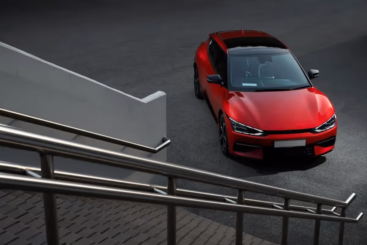 Red modern car parked on dark asphalt near metal railings and a white wall.