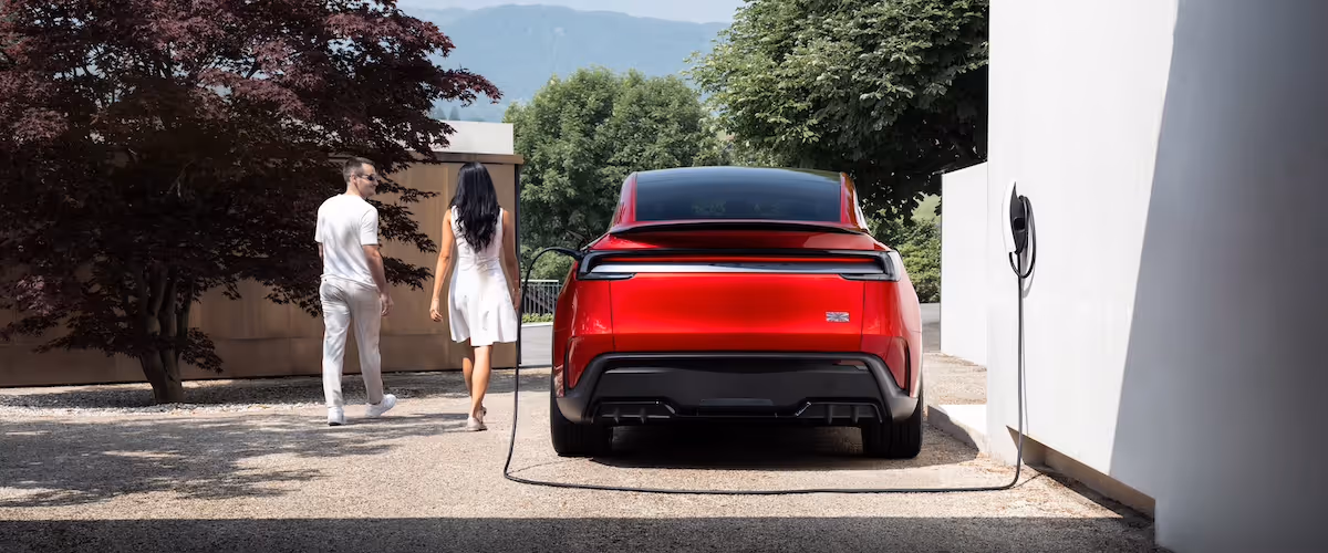 Rear view of a red electric car charging parked near a white wall with a couple walking away in a sunny outdoor setting.