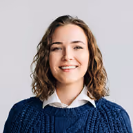 Smiling woman with curly brown hair wearing a navy blue sweater and white collared shirt.