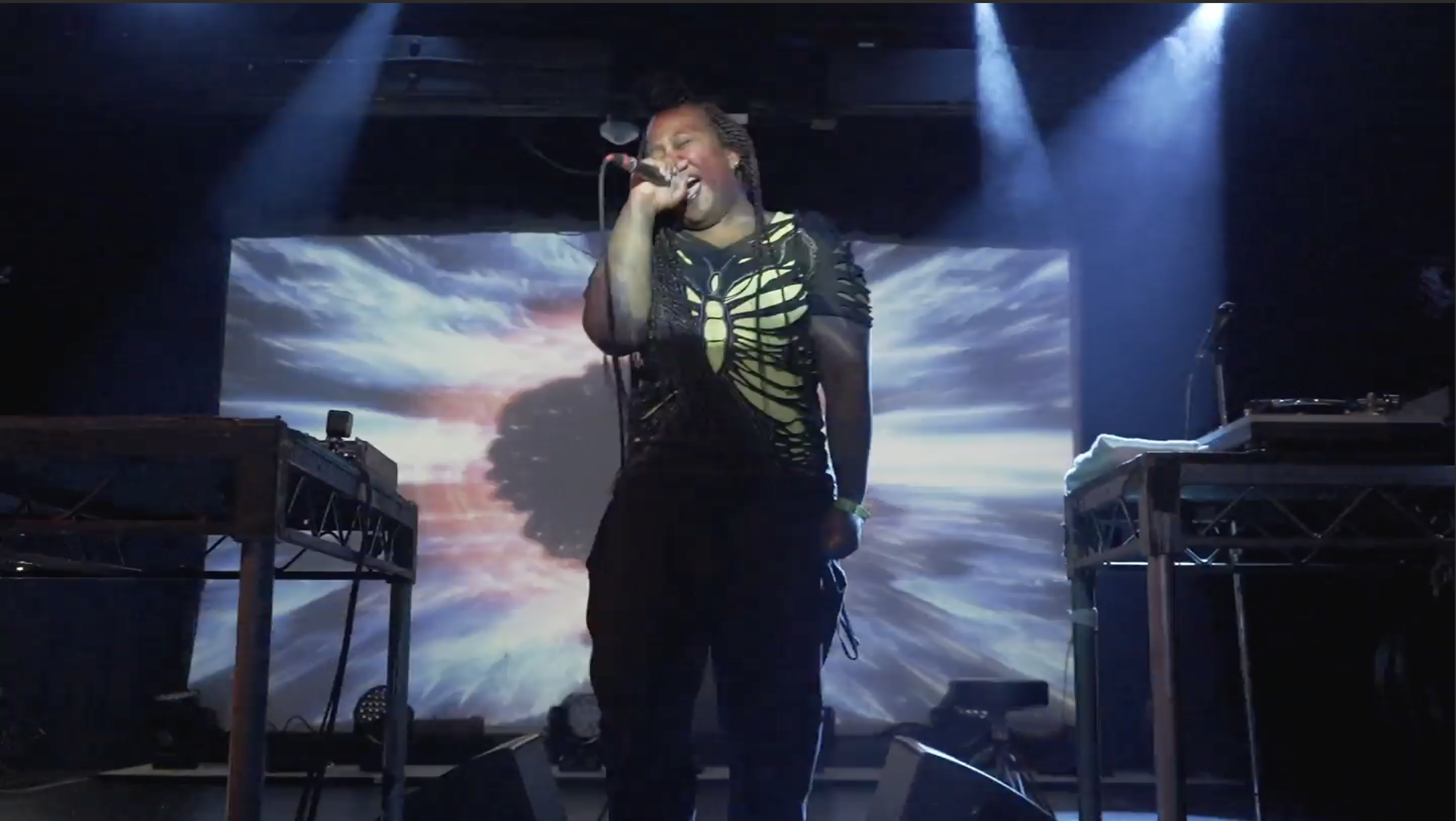 Sky Deep singing into a microphone during a live electronic music performance with immersive background visuals.