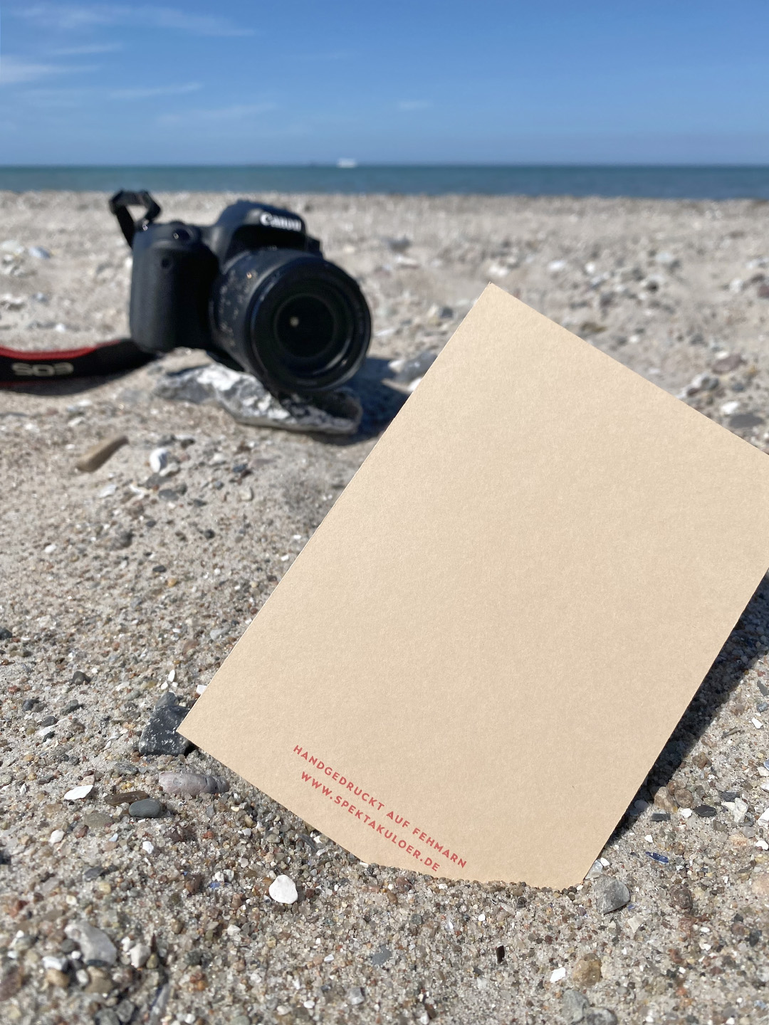 Postkarte mit dem Text »Handgedruckt auf Fehmarn« »www.spektakuloer.de« im Vordergrund, eine Kamera und Strand mit Meer im Hintergrund